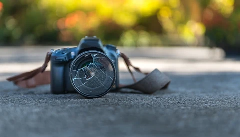 DSLR camera with cracked lens filter lying on pavement with blurred foliage background.