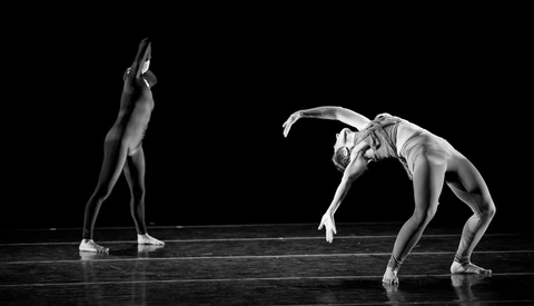 Two dancers in contrasting poses on a dark stage lit from above, one standing upright and one deeply arched backward.