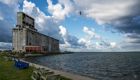 Weathered concrete grain elevator beside calm water with blue Adirondack chair on grassy shore.