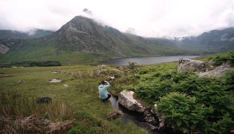 Lone hiker sitting on a rock overlooking a mountain lake surrounded by green valley and peaks.