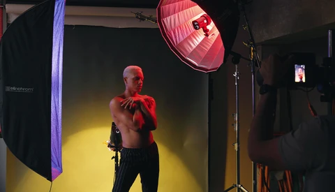 Shirtless male model posing in studio with red fabric, lit by yellow and red studio lights.