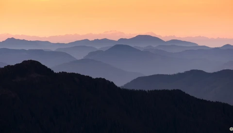 Layered mountain ridges receding into atmospheric haze at sunrise, forested slopes in silhouette.