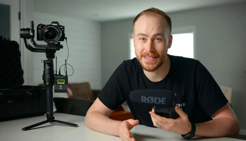 Content creator in black RODE shirt gestures while speaking to camera next to motorized gimbal with mounted DSLR.