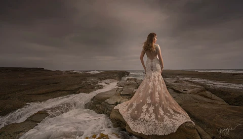 Bride in ornate lace gown standing on rocky coastal outcrop overlooking turbulent ocean.