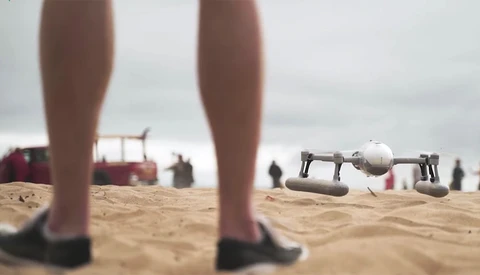 Drone hovering low over sand with person's legs in foreground on beach.