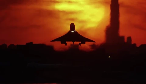 Military fighter jet silhouetted against dramatic orange and yellow sunset sky with industrial structures in background.