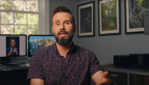 Man with beard wearing patterned short-sleeve shirt in modern office with monitors and framed artwork on walls.