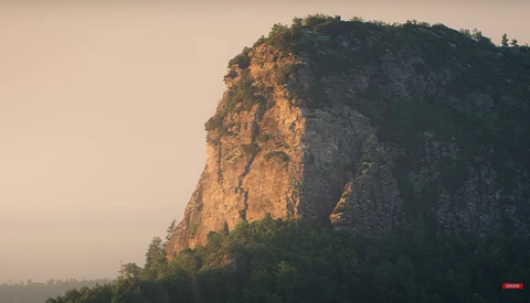 Steep rocky cliff face illuminated by golden hour sunlight with forested summit and misty valley below.