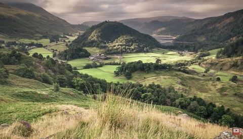 Green valley landscape with forested hills and scattered buildings under overcast sky.