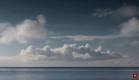 Calm seascape with puffy white clouds and blue sky above a still ocean horizon.