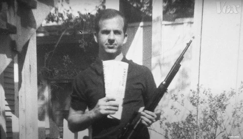 Man in dark cardigan and white shirt holding rifle, posed in front of white building with foliage.