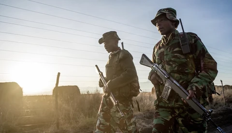Two armed soldiers in camouflage uniforms stand backlit by golden sunrise light with industrial buildings in the background.