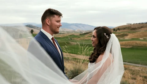 Bride and groom facing each other outdoors with veil flowing, mountains visible in background.