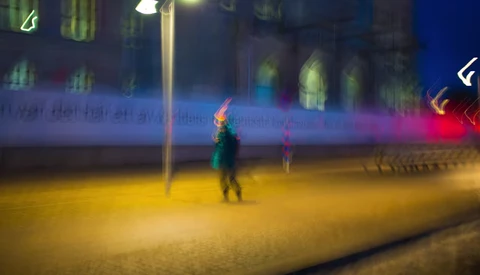 Blurred figure in green jacket standing in yellow-lit outdoor plaza at night with colorful neon signs in background.