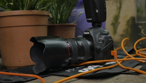Canon EOS camera with telephoto lens, external flash, and orange cables on dark surface near potted plants.