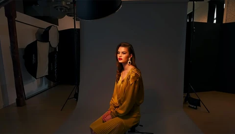 Woman in mustard-yellow dress seated in professional studio with multiple studio lights and reflectors.