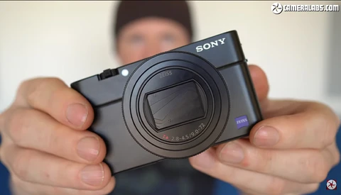 Hands holding a Sony compact camera with a zoom lens and blue Zeiss branding.