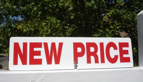 Large white sign with bold red text reading "NEW PRICE" displayed outdoors against green foliage.