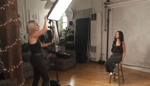 Studio portrait setup with female subject seated on stool while photographer adjusts overhead softbox lighting.