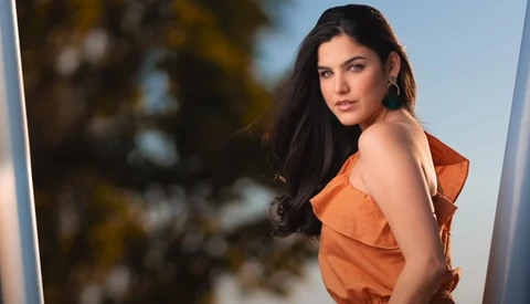 Woman with long dark hair wearing an orange sleeveless top, posed against a blurred natural background.