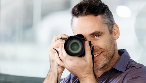 Male photographer holding a DSLR camera up to his face, smiling while composing a shot in a bright indoor setting.