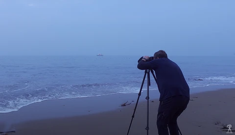 Photographer using tripod and telephoto lens on a beach at dusk, shooting toward the ocean horizon.