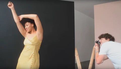 Photographer shooting a portrait of a woman in a yellow dress posed against a dark backdrop in a studio setting.