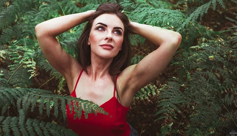 Woman in red halter top posing among lush green ferns with hands behind head.