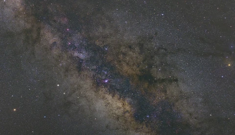 The Milky Way galaxy stretches across the night sky with vibrant purple and blue nebula regions amid dense star fields and dark dust lanes.