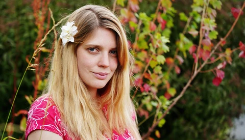 Young woman with long blonde hair and white flower accessory, wearing pink patterned top against autumn foliage background.