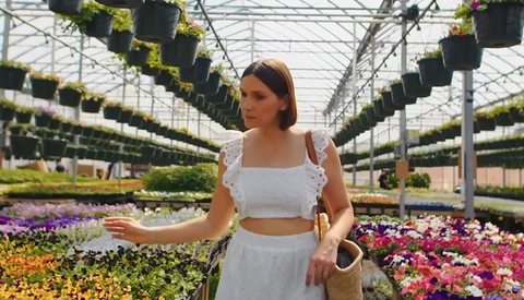 Woman in white crop top and skirt standing in greenhouse surrounded by colorful flowering plants.