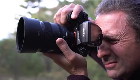 Photographer holding a mirrorless camera with telephoto lens to his eye while shooting outdoors.