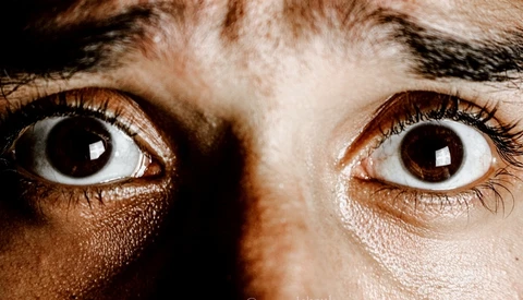 Close-up of wide-eyed human eyes with brown irises against dark background, showing intense expression.