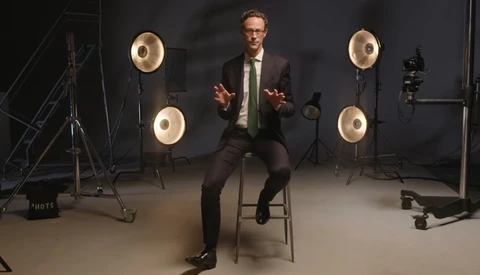 Man in business attire sitting on a stool in a professional studio setup with ring lights.