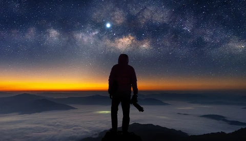 Silhouetted figure standing on a mountain peak overlooking a sea of clouds at sunset with the Milky Way visible above.