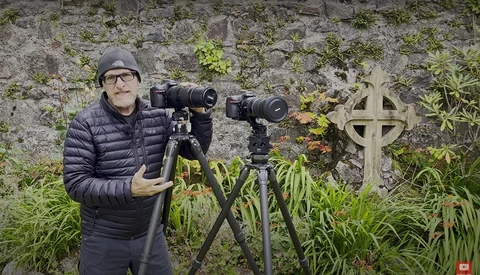 Photographer with telephoto lenses on tripods in front of moss-covered stone wall and cross.