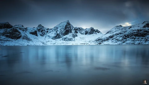 Snow-covered mountain peaks reflected in a still, partially frozen alpine lake at twilight.