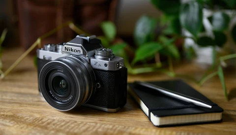 Nikon Z camera with attached lens on wooden desk beside closed notebook, indoor plants visible in background.