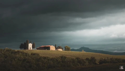 Rural chapel and farmhouse on a hillside under dramatic storm clouds.