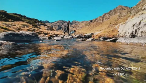 Shallow mountain stream flowing over rocky bottom with hikers standing in distant valley under clear sky.