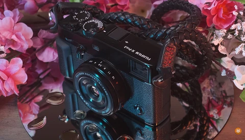 Black rangefinder camera with blue accents displayed on a reflective surface surrounded by pink and red flowers.