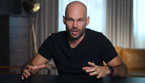 Man in black v-neck t-shirt gestures while speaking during an interview or presentation in a modern interior.