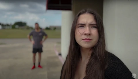 Young woman with long dark hair looking toward camera while person exercises in blurred background.