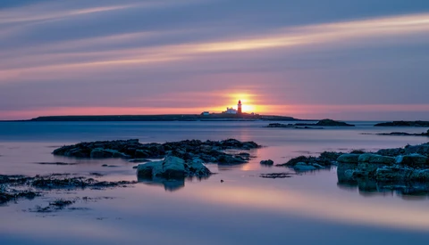 Lighthouse on distant island at sunset with rocky foreground and calm water.