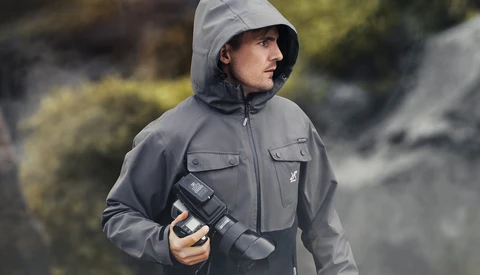 Photographer wearing gray technical jacket and hood while holding a telephoto lens outdoors.