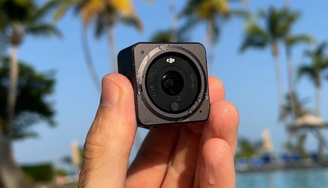 DJI action camera held in hand against tropical waterfront with palm trees and blue sky.