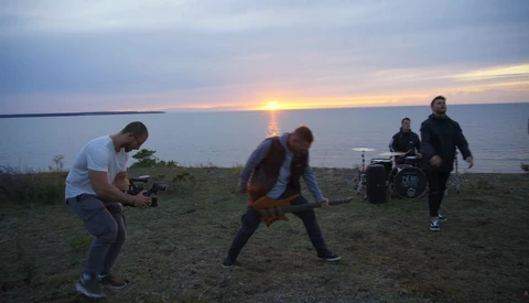 Musicians performing on a grassy beach overlooking water at sunset, with a drummer and guitarist visible.