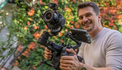 Filmmaker holding a cinema camera rig with mounted gimbal stabilizer against a flowering vine backdrop.