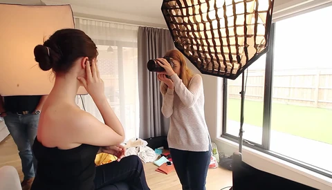Photographer adjusting a softbox light modifier while model sits during indoor studio portrait session.