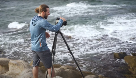 Photographer on rocky shore operating camera mounted on tripod, facing ocean waves.